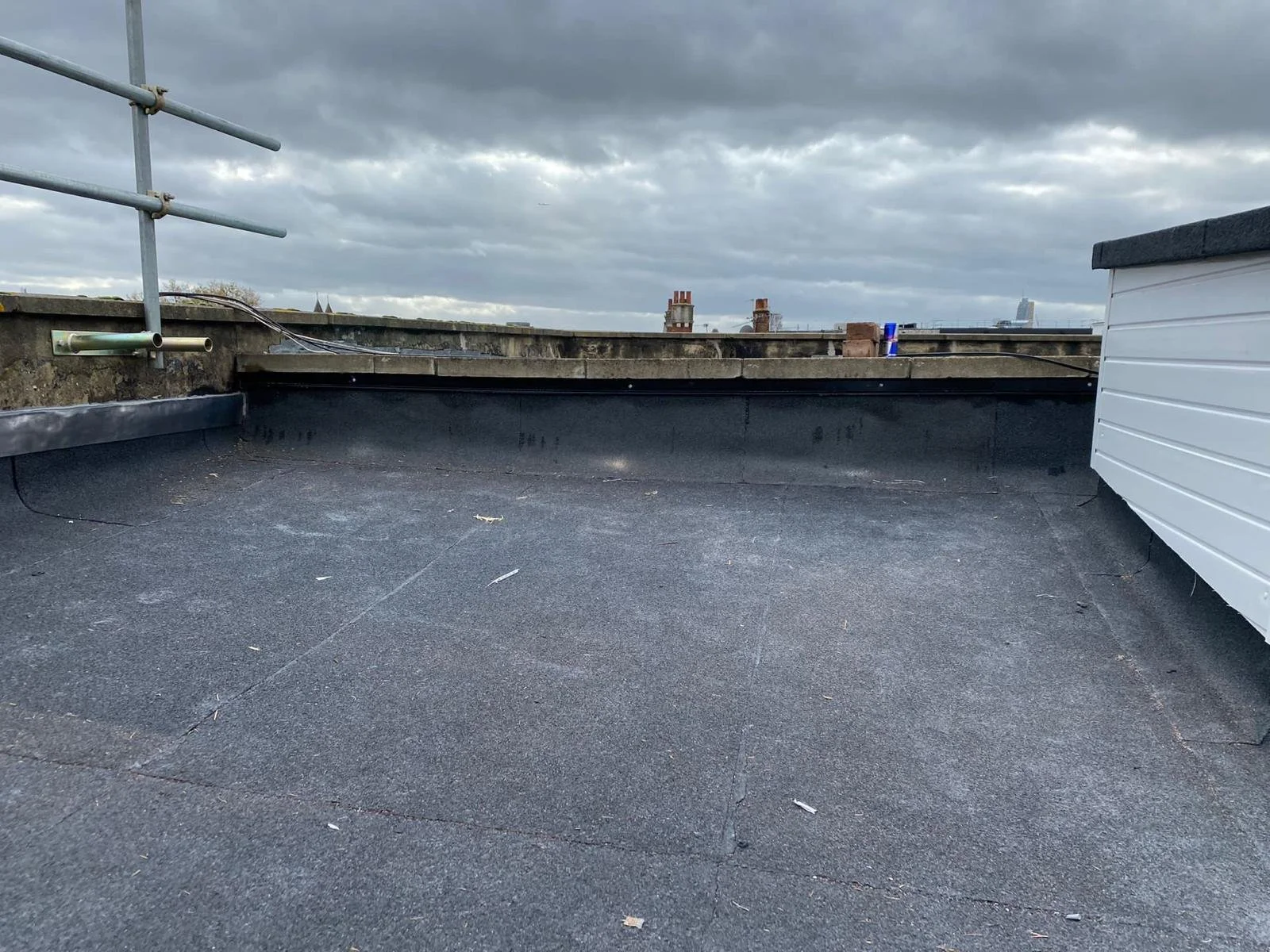 On a rooftop, grey asphalt surface with a low brick wall and metal scaffolding on the left, and cloudy sky overhead with some distant buildings visible.