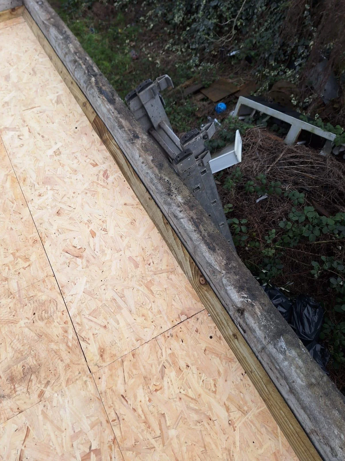View from a construction site showing plywood flooring, a wooden beam, and a power nailer tool attached to the beam. In the background, there is a garden with some discarded items including a white chair, a blue plastic bottle, and debris.