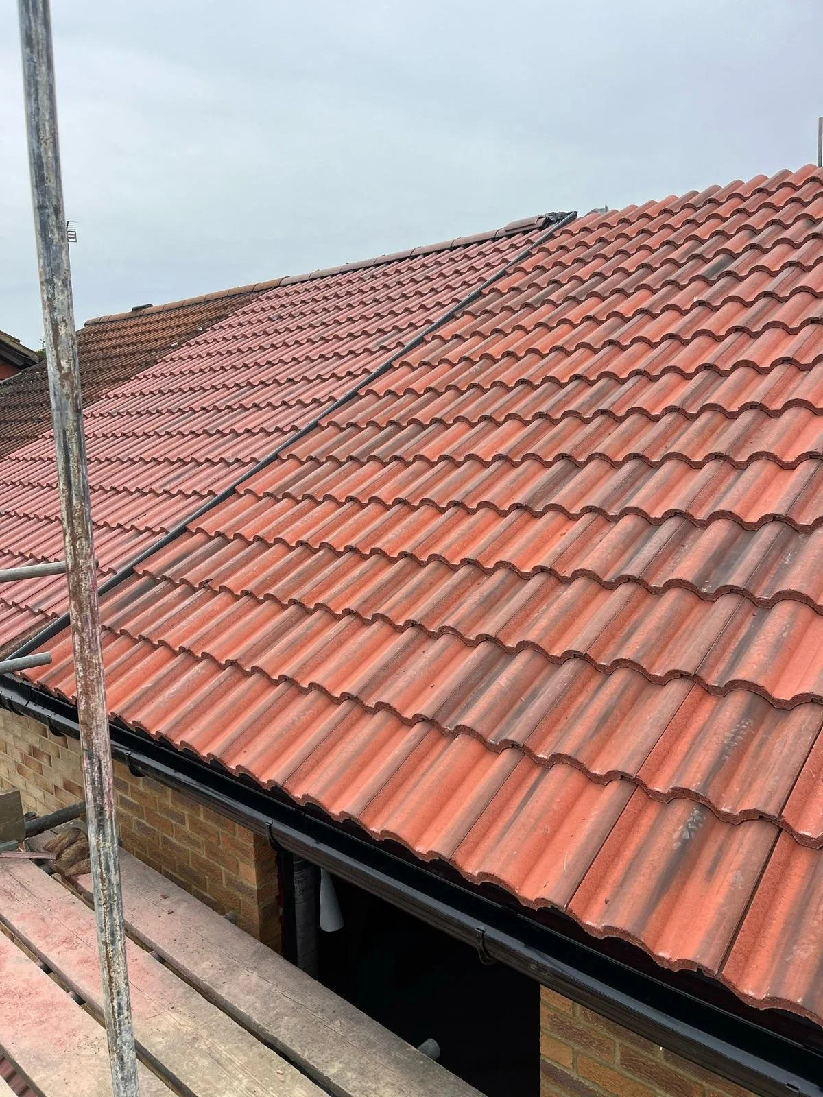 Close-up of a house roof with reddish-orange tiles under an overcast sky, with scaffolding and brick wall visible at the edge.