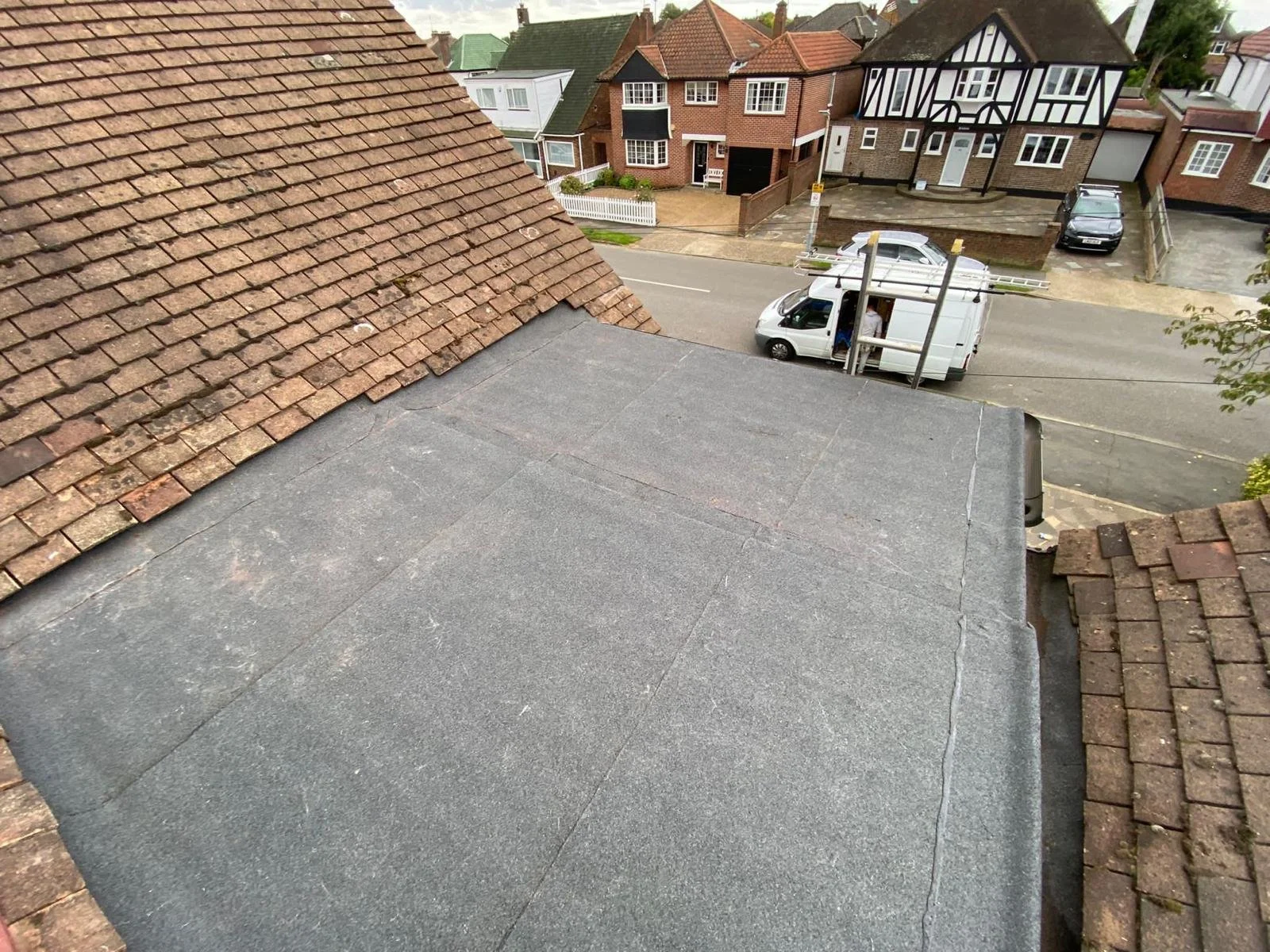 A view from a roof showing a quiet residential street with parked cars, a delivery van, and houses with varied architecture including a traditional half-timbered house.