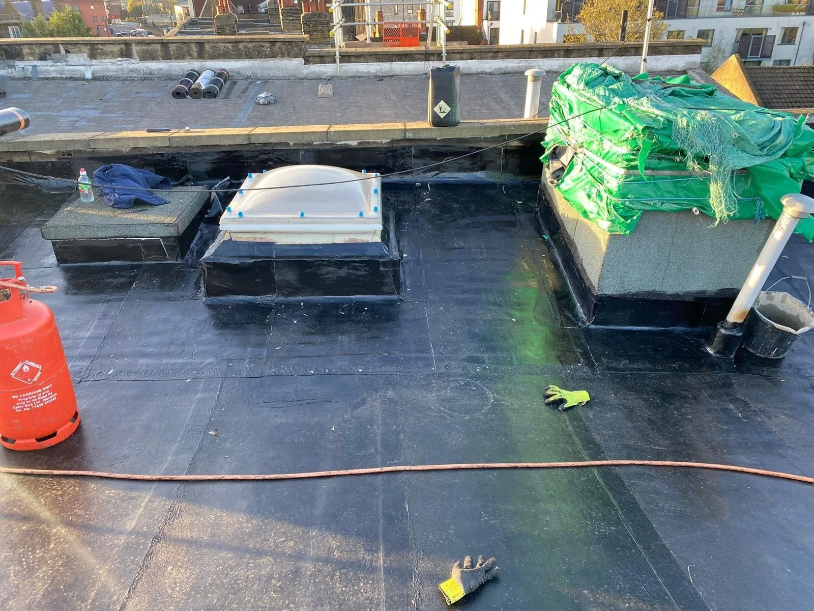Rooftop with construction materials and tools, including a propane tank, gloves, green tarp, pipes, and small ventilation units, with neighboring buildings visible in the background.