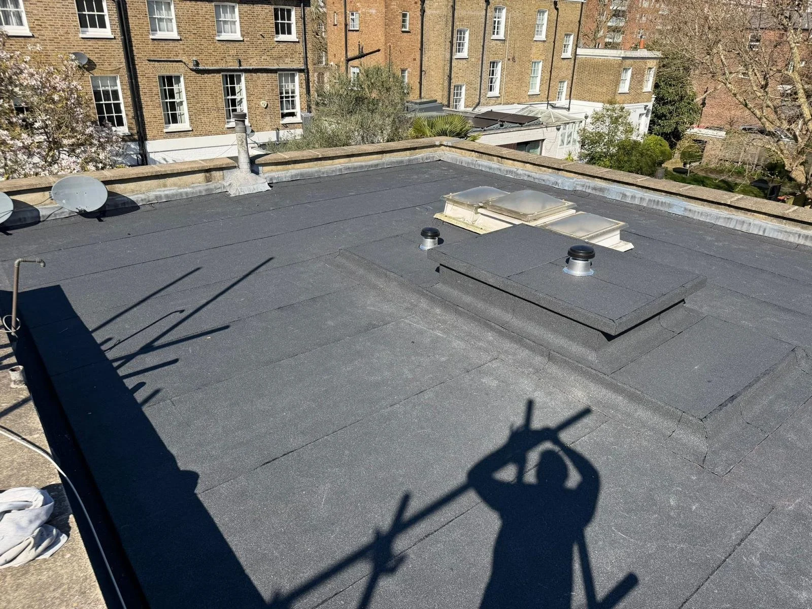 Rooftop with black tar paper, vents, and skylights, surrounded by brick buildings and trees with budding leaves, shadows of the photographer and a pole are cast on the roof.