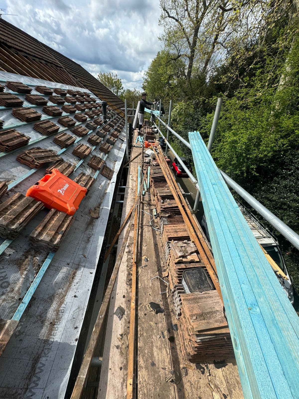 A person working on a roof undergoing repairs, with roofing tiles and construction materials present, on a scaffold next to a building with trees in the background.