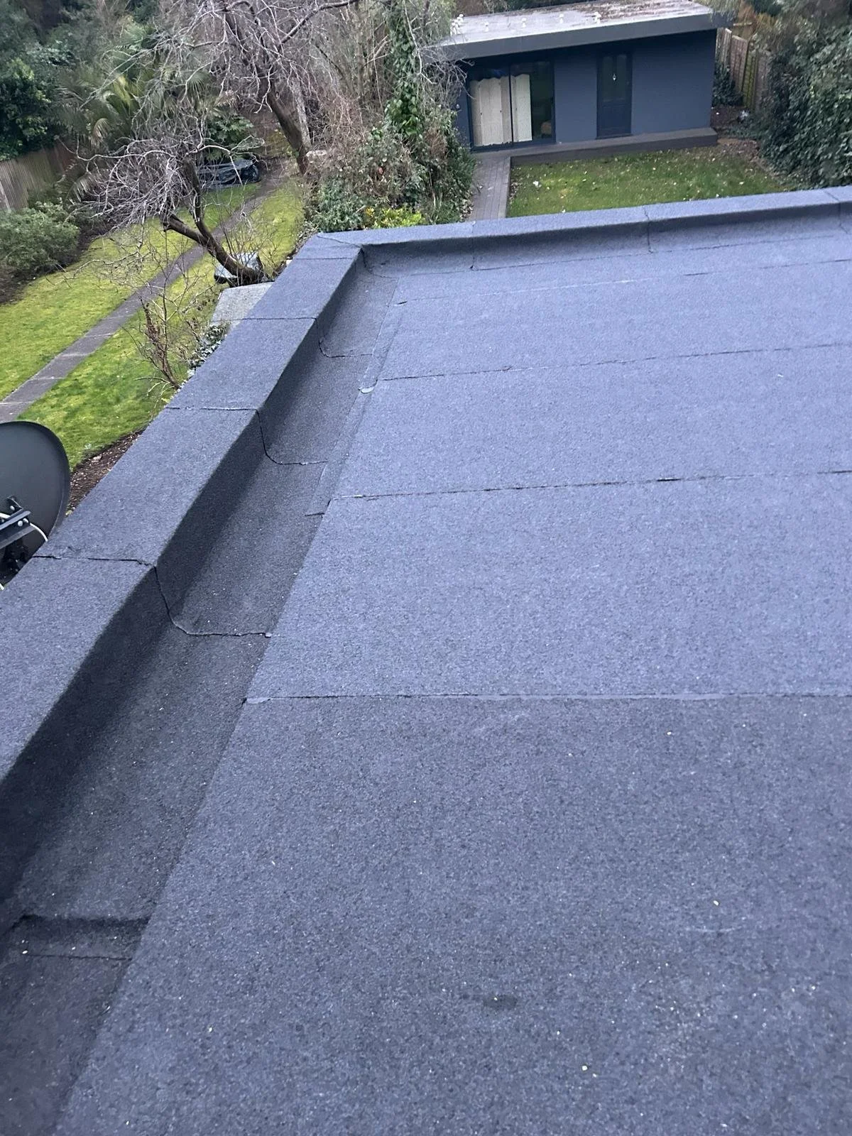 View of a flat roof with dark shingles, showing a slight inclination at the edges and a cracked corner, overlooking a backyard with a small structure, trees, and green grass.