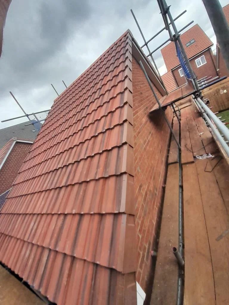 Close-up of a brick chimney with new red clay roof tiles installed, surrounded by scaffolding on a house roof under a cloudy sky.