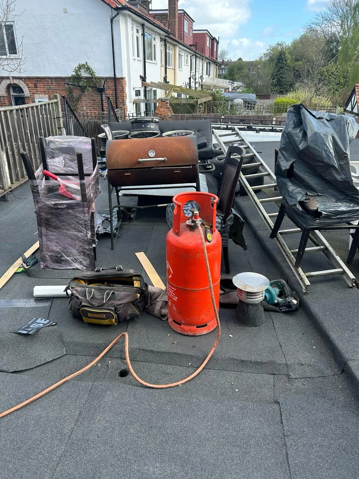 Construction site on a rooftop with tools, a propane tank, ladder, and furniture, amid building supplies and a residential neighborhood background.