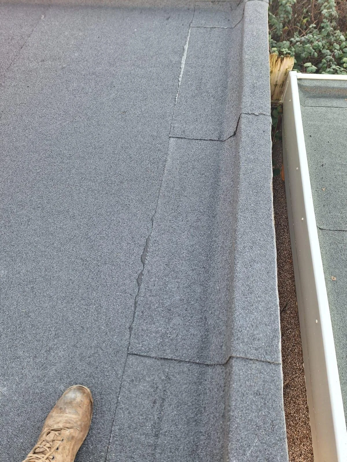 Close-up of a rooftop with gray asphalt shingles, showing a crack and some lifted shingles near the edge. A person's boot is visible at the bottom of the image.