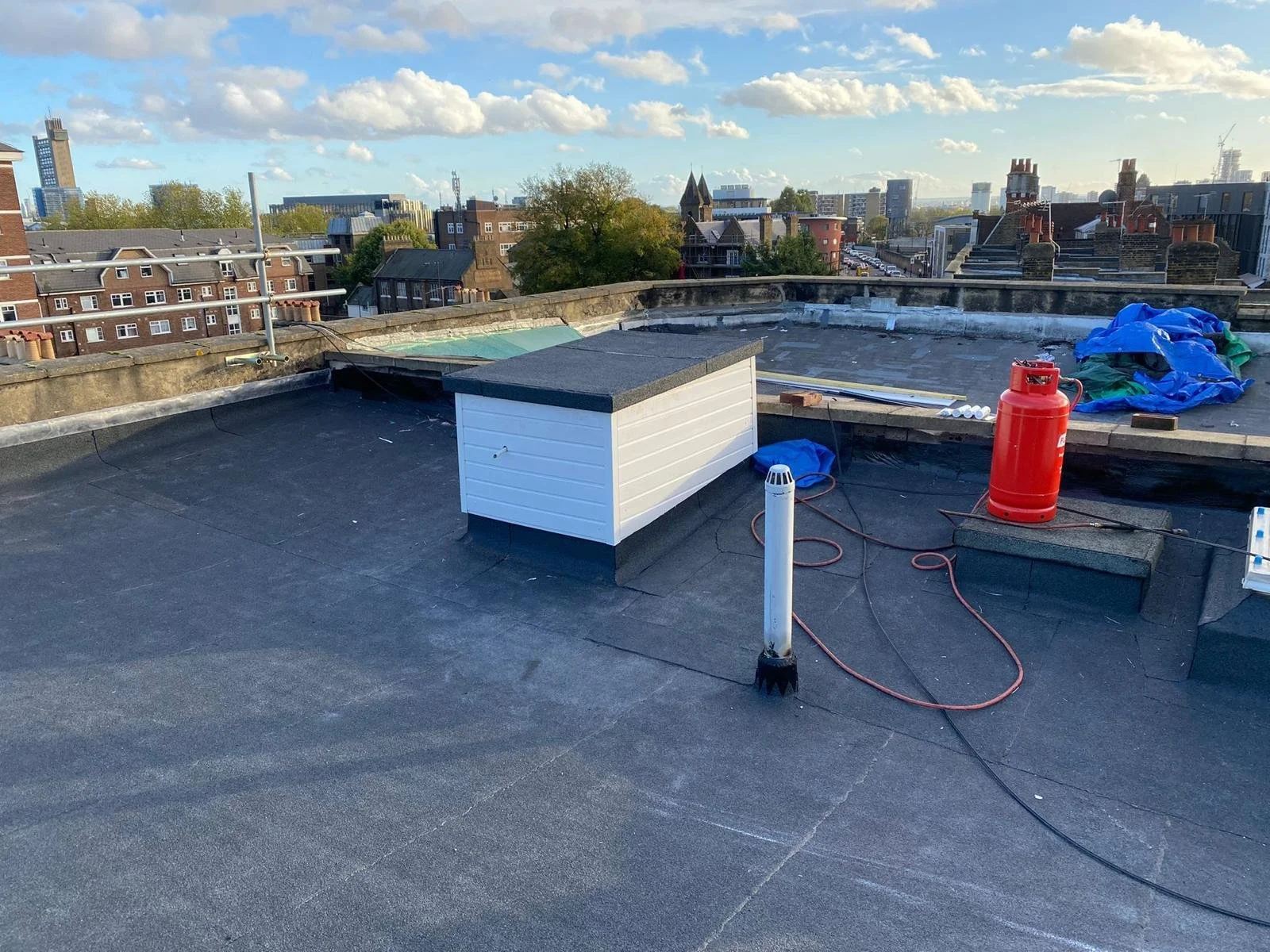 View of a rooftop with construction or maintenance equipment, including a red propane tank, a white vent pipe, and a small white shed, with a cityscape and partly cloudy sky in the background.