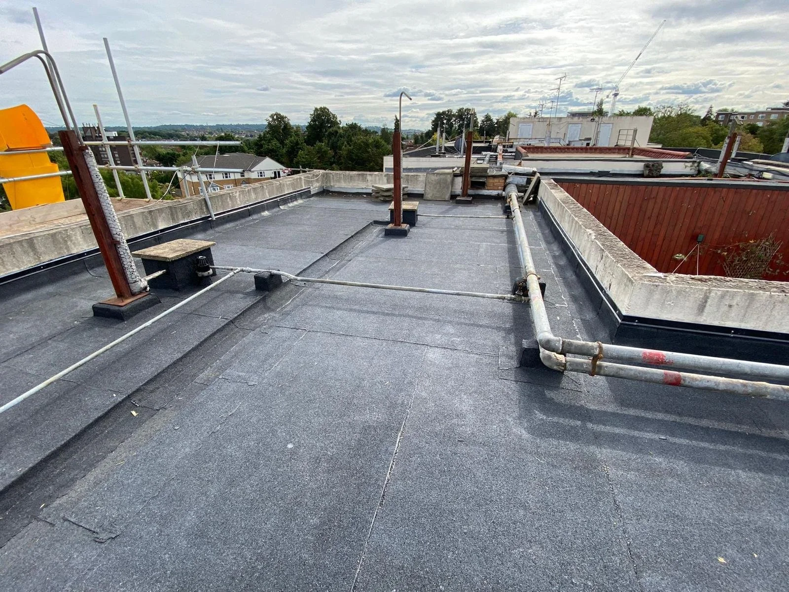 Flat rooftop with black roofing material, pipes, vents, and a few small structures, surrounded by a parapet wall, with other rooftops and trees visible in the background under a cloudy sky.