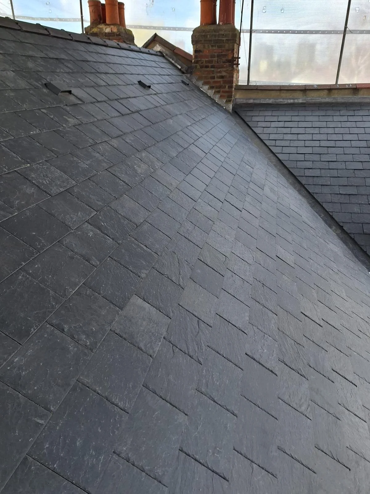 Close-up of a slate roof, showing the textured, overlapping shingles and part of a brick chimney with two pipes.