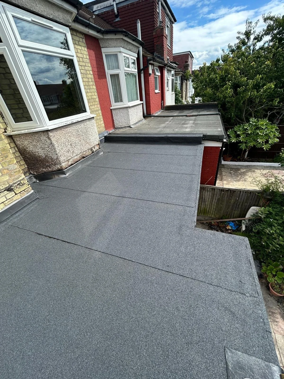 View of a flat roof with grey waterproofing material on a row of houses, showing part of the houses' brick walls, windows, and a tree on the side.
