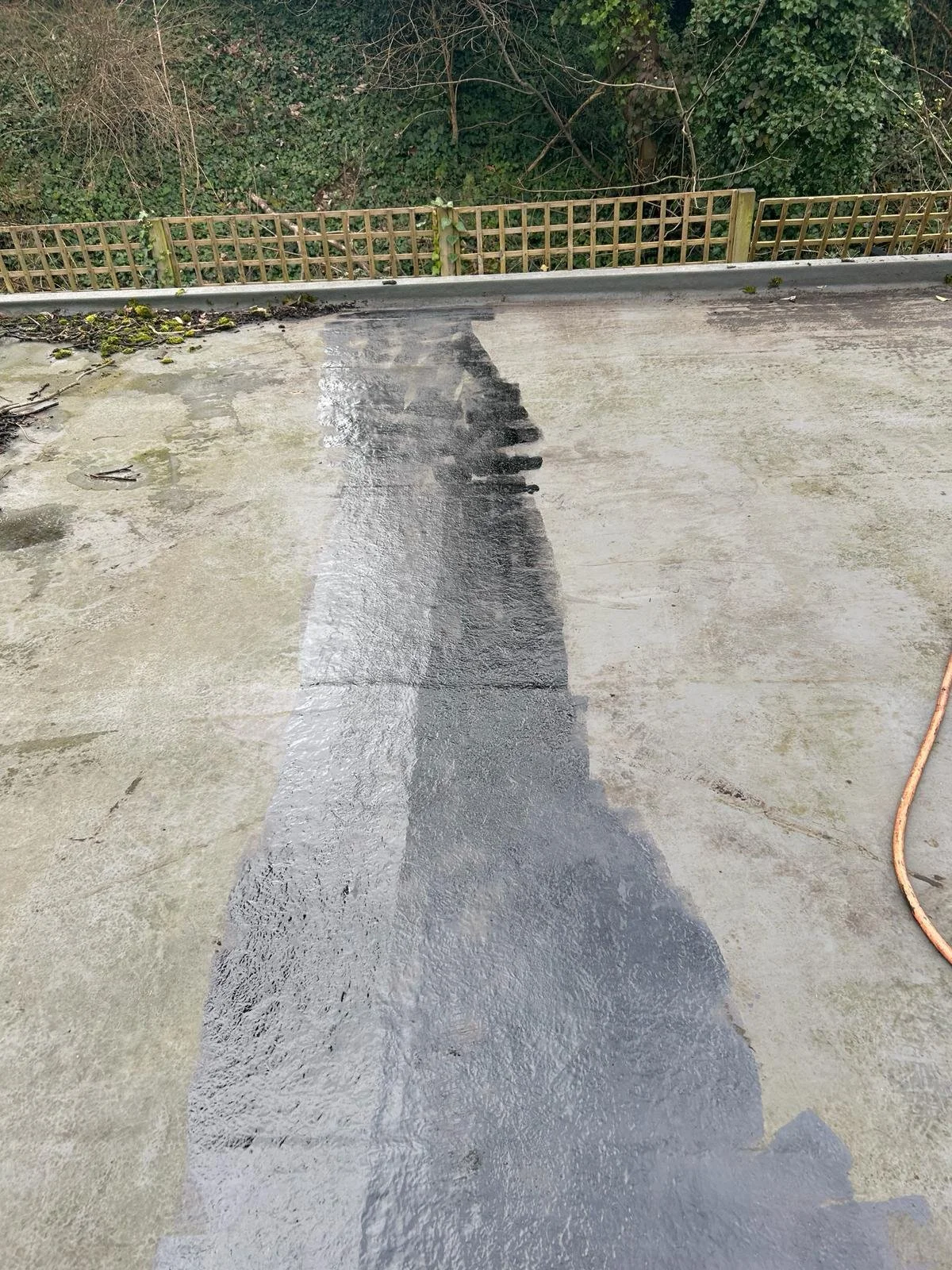 A concrete surface with a black waterproofing coating partially applied, with a hose visible on the right side and a wooden fence in the background.