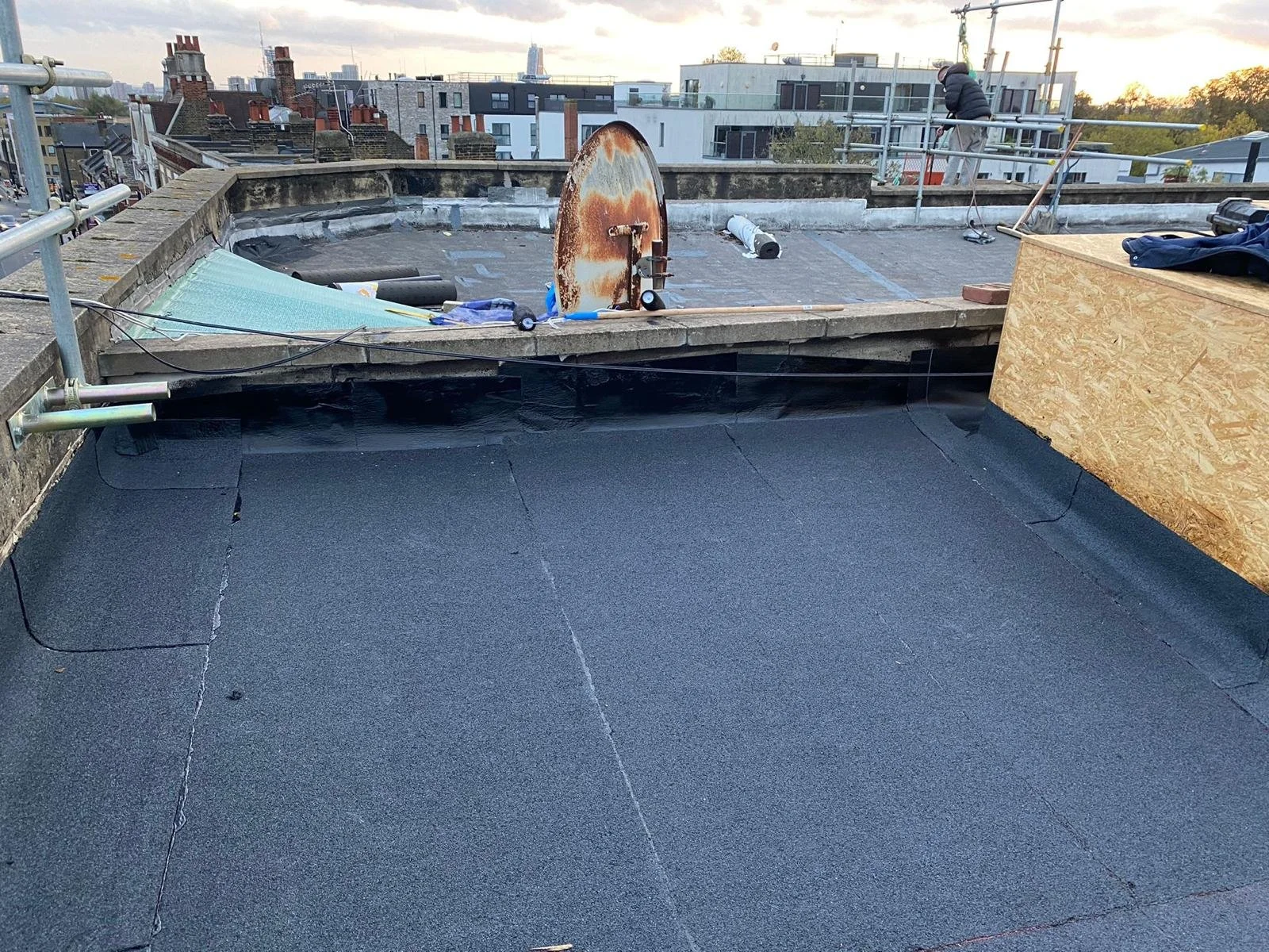 View of a rooftop under construction, with black roofing material, scaffolding, and construction tools, and a cityscape in the background during sunset.