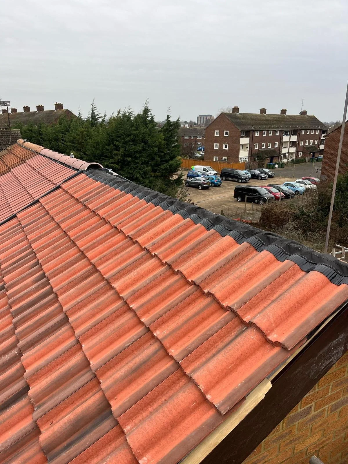 View from a rooftop showing orange and gray tiled roof, trees, and a parking lot with cars and apartment buildings in the background.