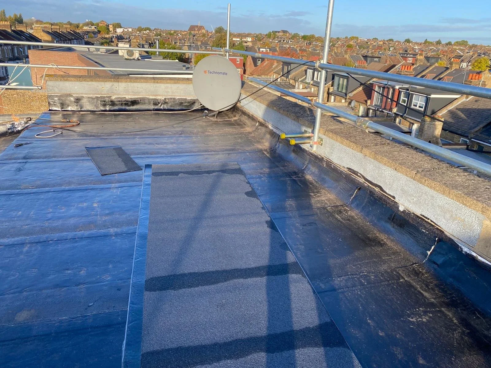 Flat rooftop with black waterproofing membrane, satellite dish, and metal railing, overlooking a neighborhood of rooftops under a clear sky.