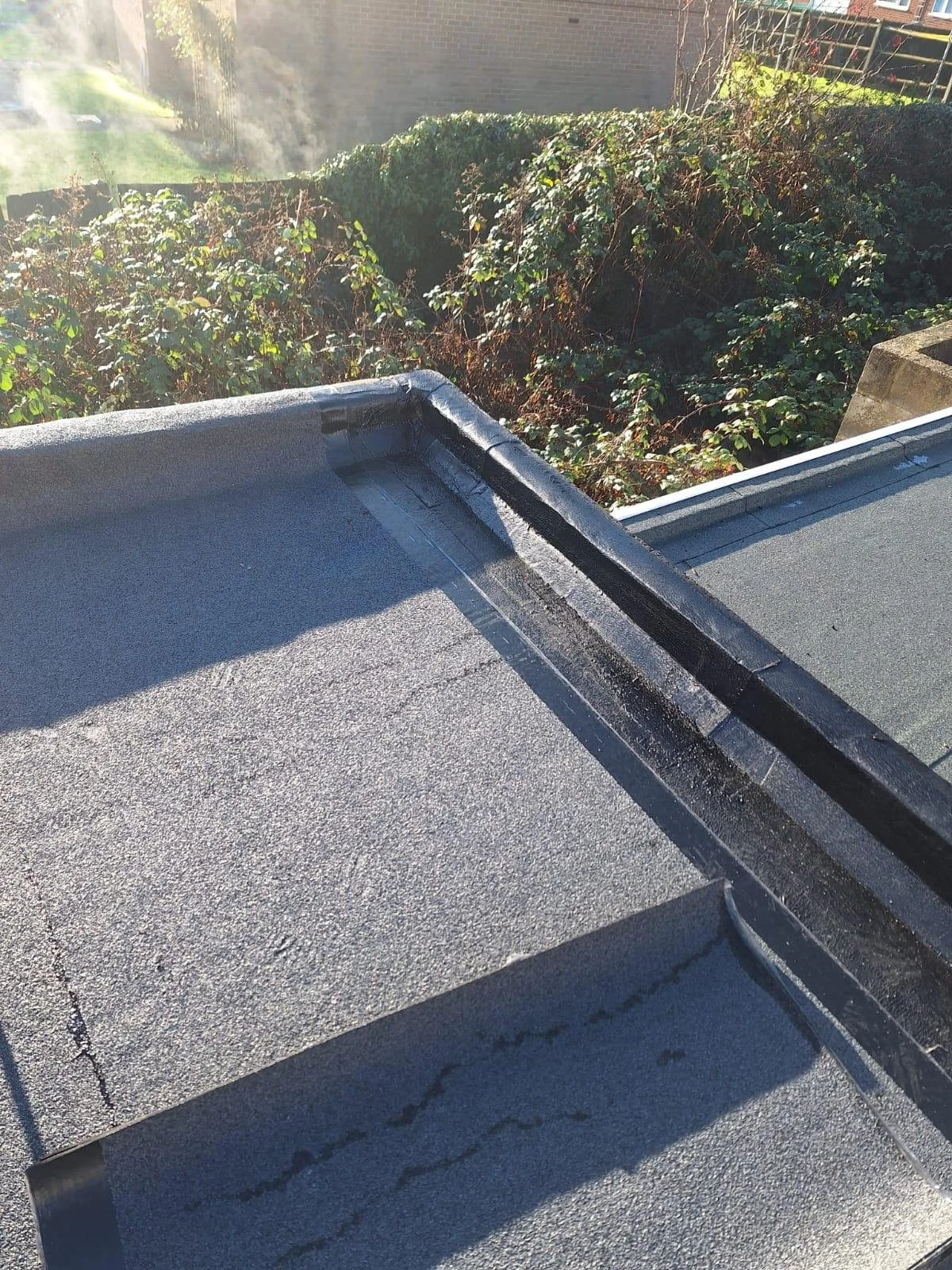 Close-up of a flat roof Maida Vale with black flashing and gravel surface, with greenery and sunlight in the background.