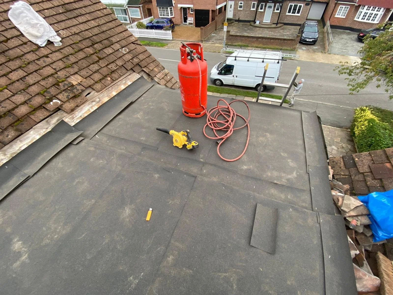 View from a roof showing construction tools and materials, including a red gas cylinder, a yellow cordless drill, a coiled hose, and some roofing tiles.