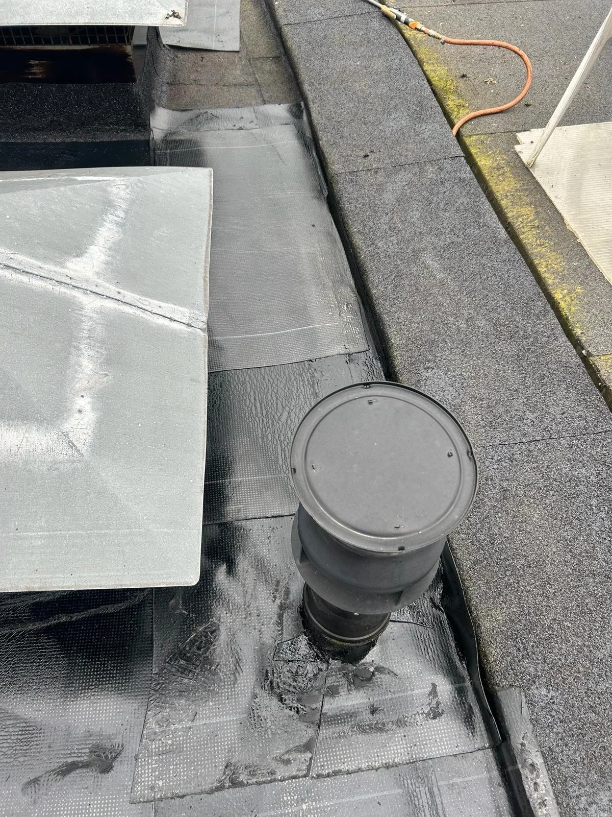 Close-up of a flat rooftop with metal flashing, a small black vent pipe, and a cable on the edge of the roof. The roof surface is covered with black waterproof material, and there is a gravel area with a white pole.