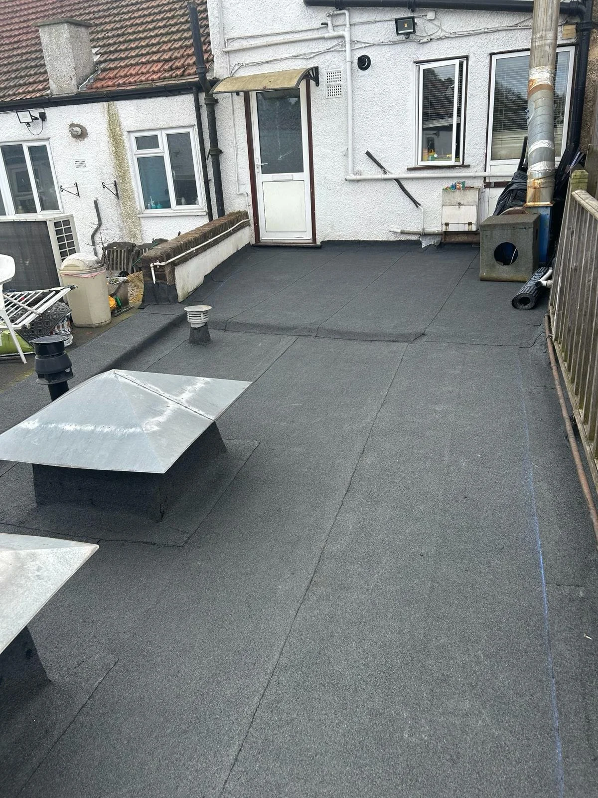 Flat rooftop with black roofing material, two ventilation vents, and a stainless steel skylight. A door leads to the interior, and there are windows, pipes, and various objects around the edges of the roof.