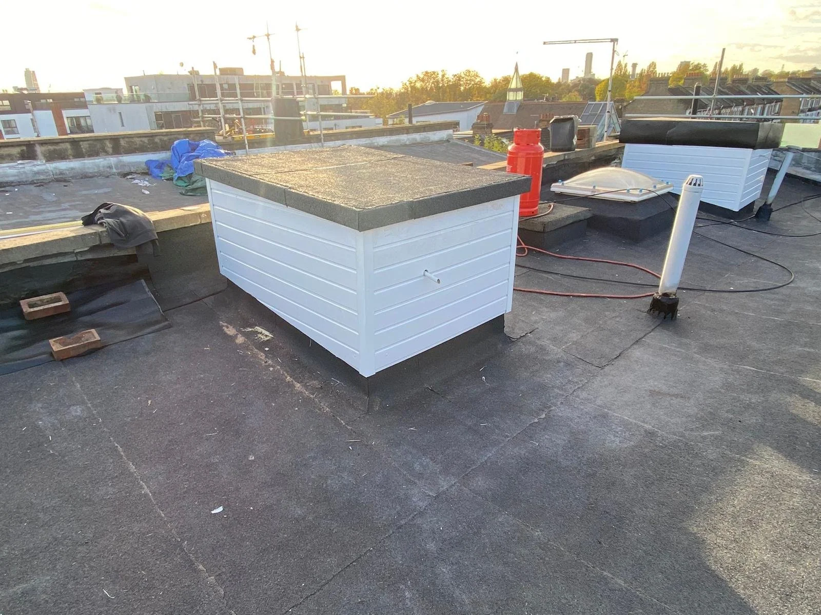 Roof with various rooftop equipment, including a white vent pipe, a red fire extinguisher, and several white rooftop structures, with city buildings and cranes in the background at sunset.
