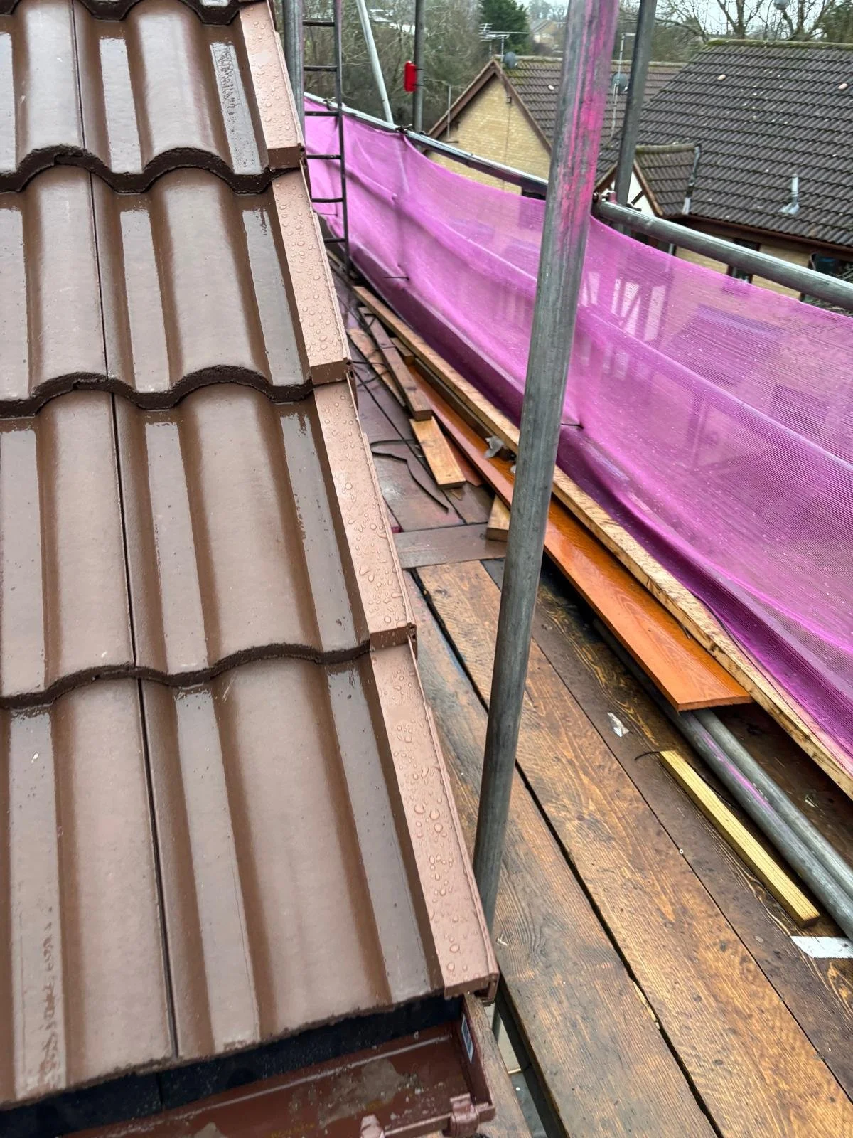 View from a roof construction site showing brown roof tiles, scaffolding with pink safety net, and neighboring houses in the background.