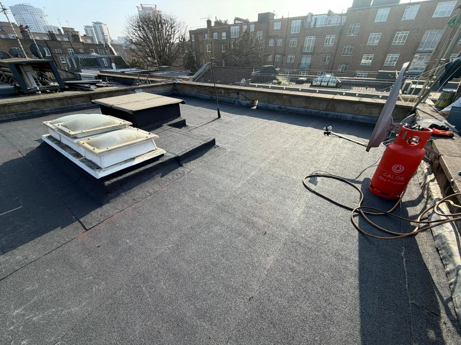 Roof with black felt and HVAC vents, a propane tank, a solar panel, and various equipment with urban buildings in the background.