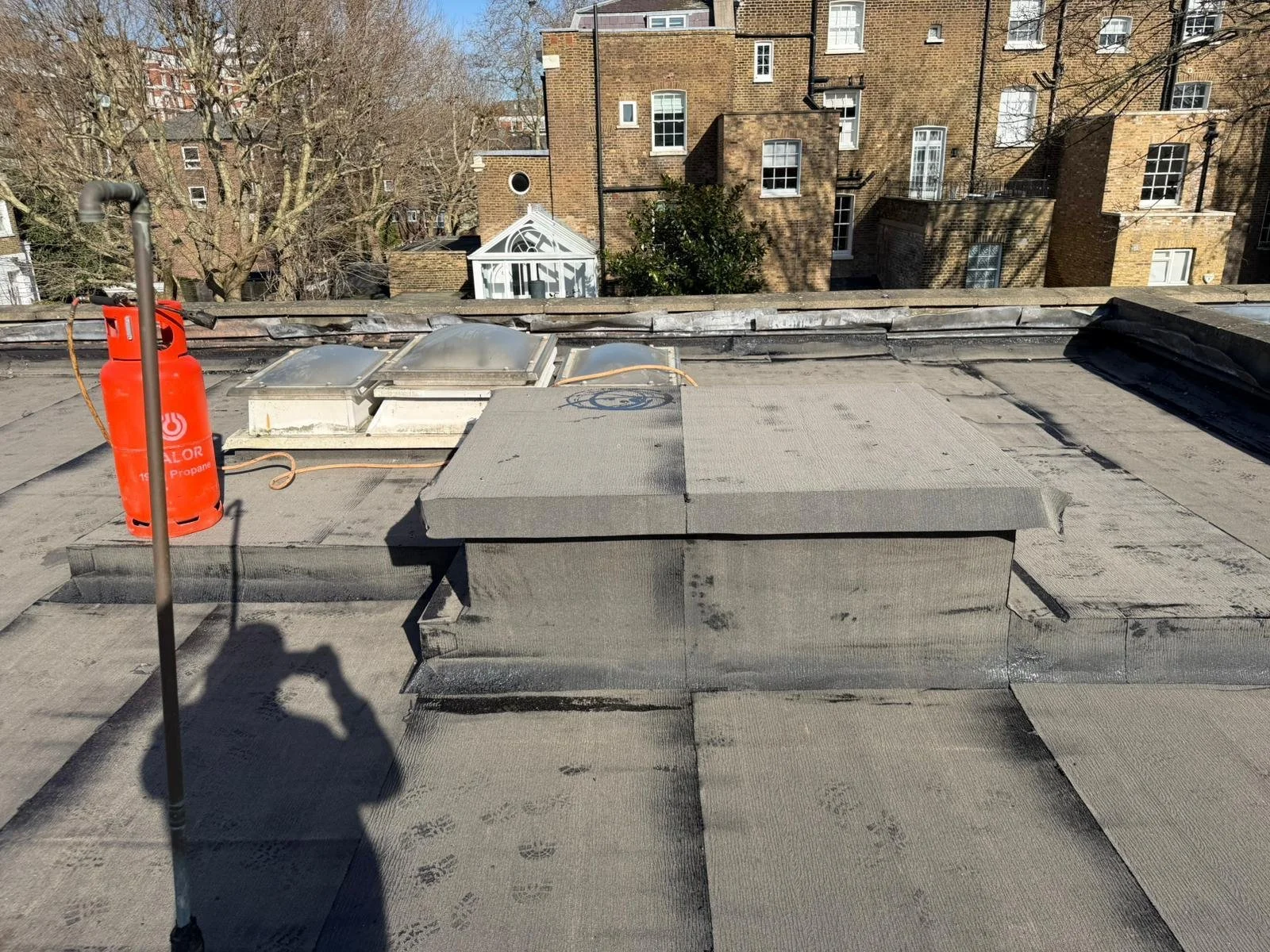 Rooftop with a red propane tank, few skylights, a small glass greenhouse, and surrounding brick buildings visible in the background.
