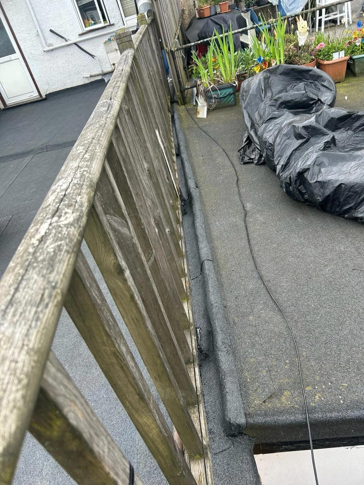 A rooftop deck with a wooden railing, potted plants, a black plastic water cover, and a black tarp covering some objects, with a house wall in the background.