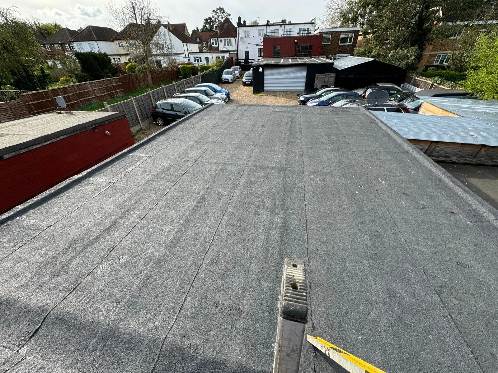 View from the rooftop of a building showing a parking lot with several parked cars, surrounded by fences and nearby houses with trees and cloudy sky in the background.