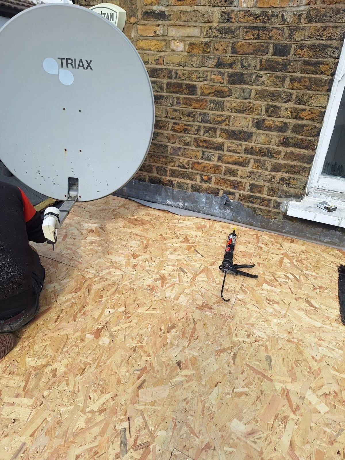 Construction area with OSB plywood flooring, a satellite dish labeled 'TRIAX', a caulking gun, and a brick wall with a window.