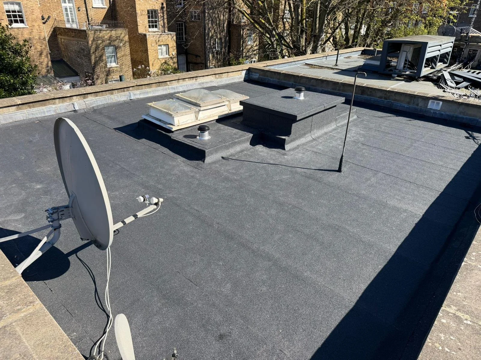 View of a flat rooftop with two skylights, two small vents, a satellite dish, and a satellite dish cable, overlooking neighboring brick buildings and trees.