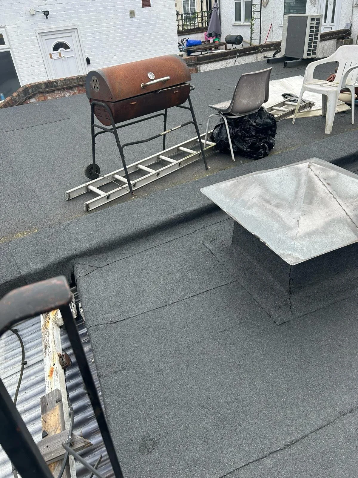 On a rooftop, there are a rusty grill, a ladder, two plastic chairs, a trash bag, and some miscellaneous items near the building's exterior wall.