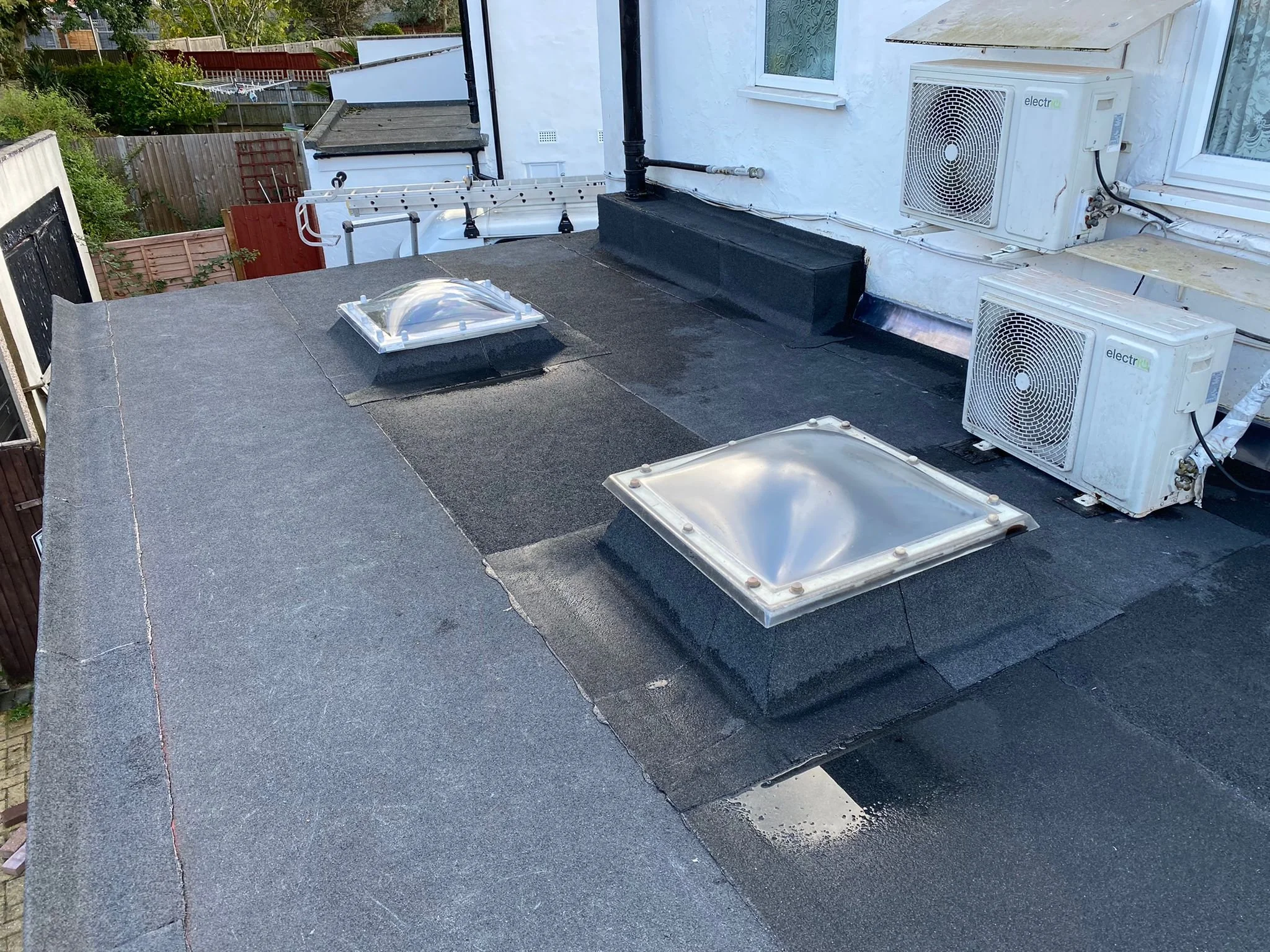 View of a flat roof with two metal skylights, in a residential area. There are two air conditioning units mounted on the wall. The roof surface appears to be covered with dark roofing material.