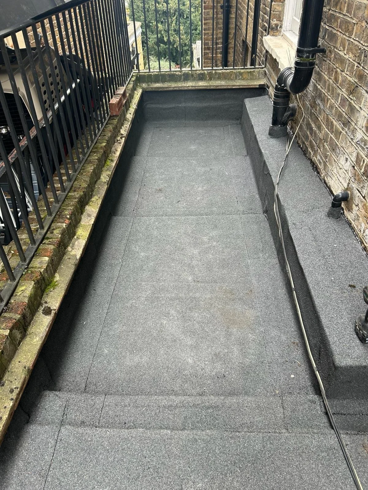 Small balcony with black metal railing, waterproof flooring, brick wall, and visible plumbing pipes.