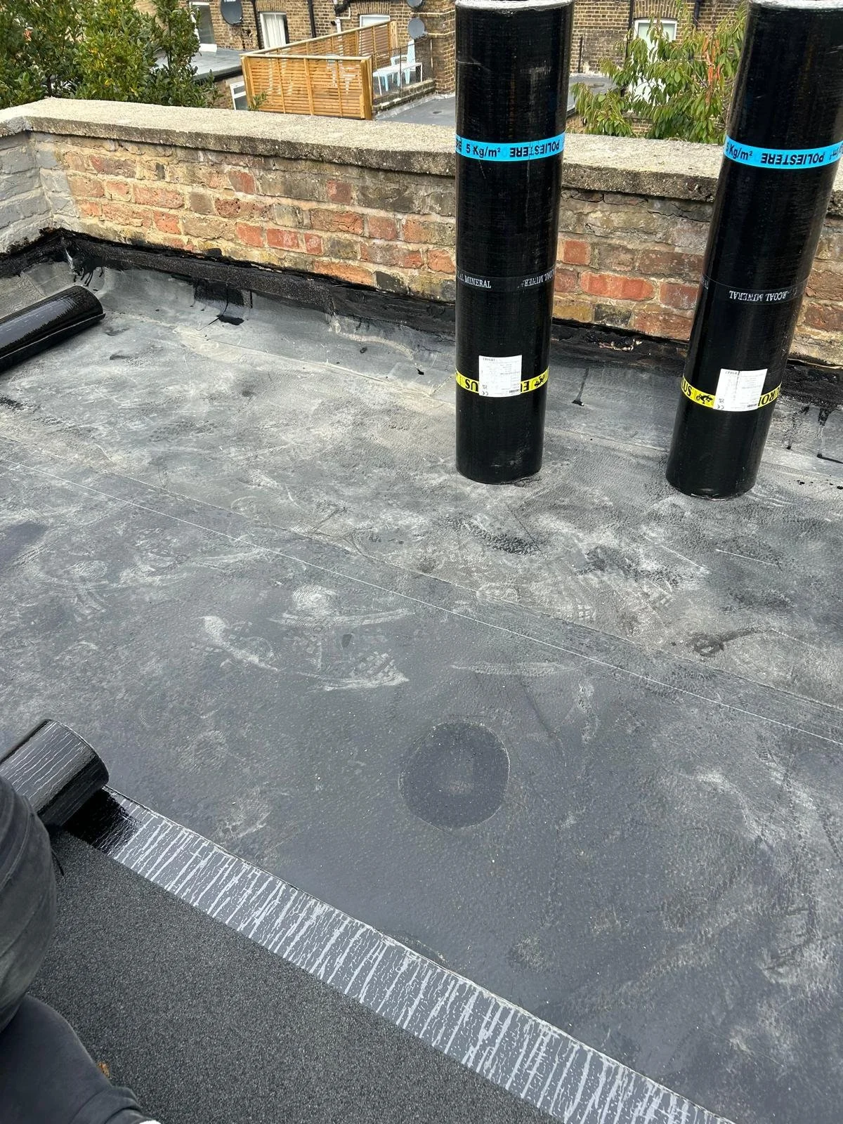 Roof with two black ventilation pipes wrapped with blue and yellow labels, a brick parapet, and a flat surface with visible footprints and a layer of roofing material.