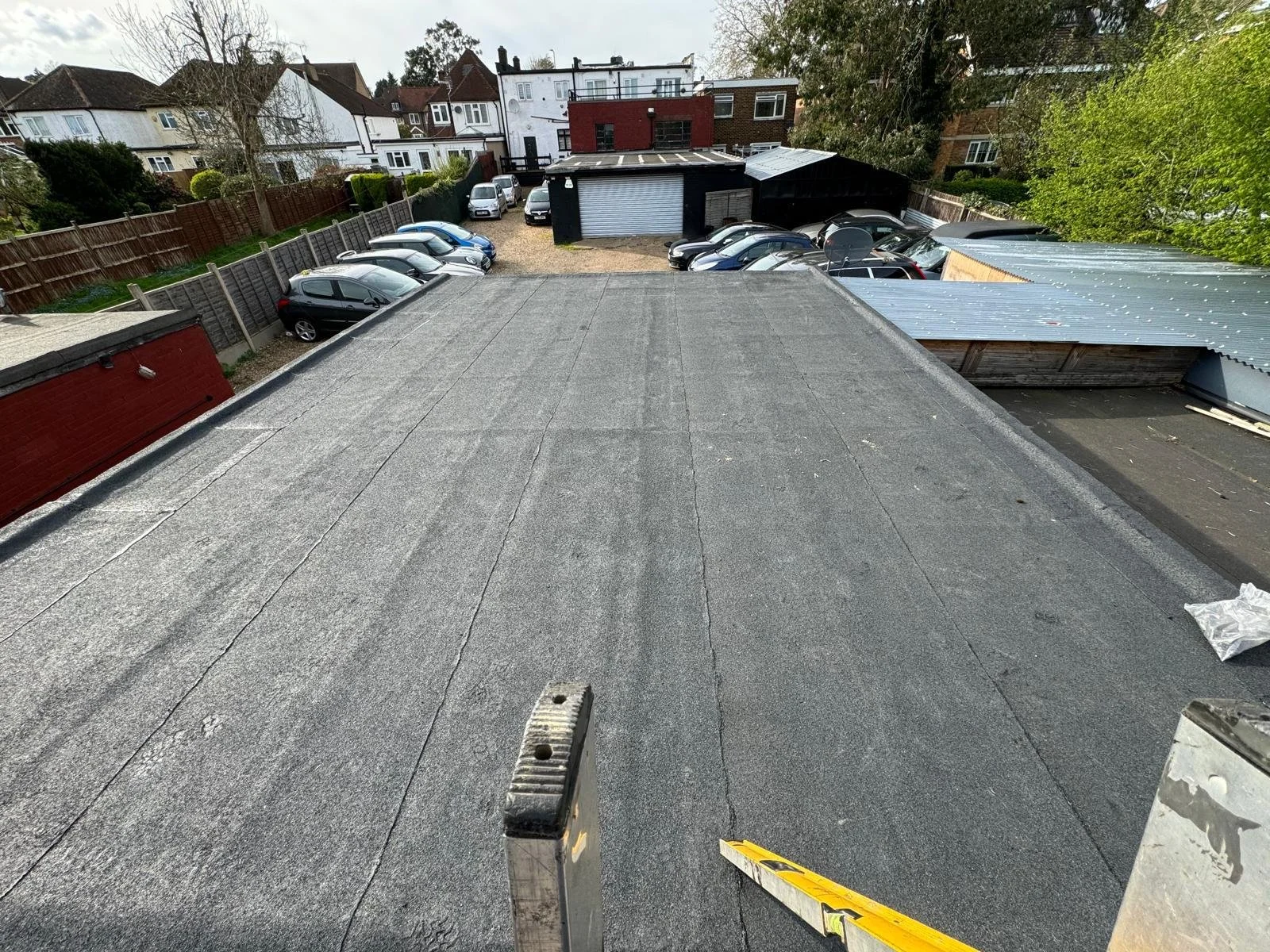 View from a rooftop showing a flat roof surface, a ladder, and construction tools, with a parking lot and residential houses in the background.