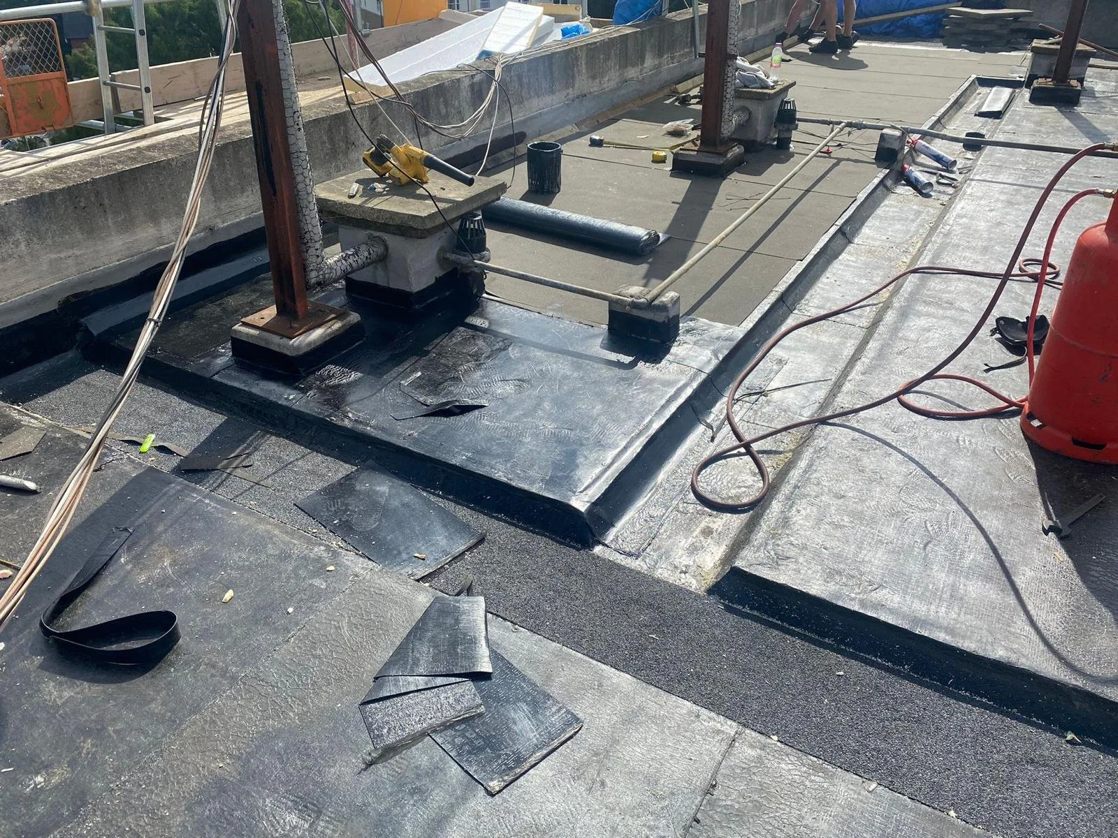 Construction workers installing or repairing a flat roof, with roofing materials and tools visible, including a propane tank and sealing patches.