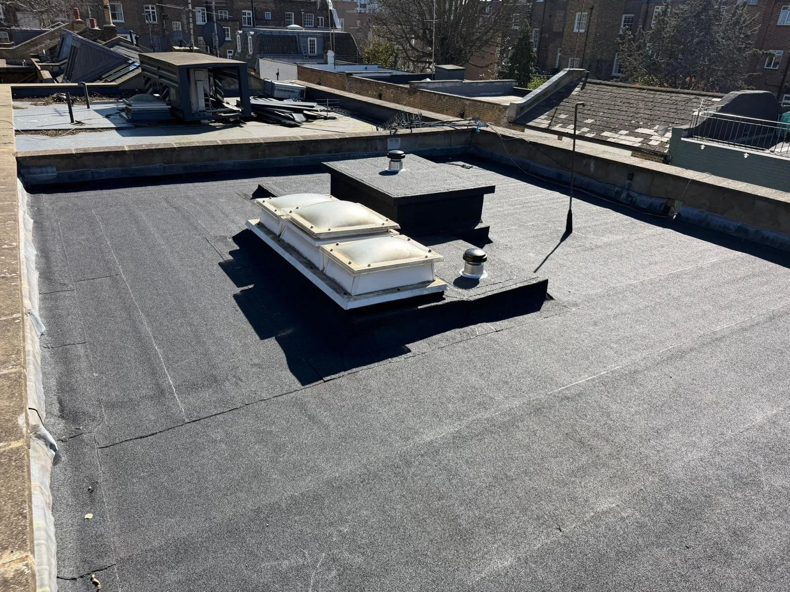 View of a flat rooftop with black roofing material, roof vents, skylights, and surrounding buildings.