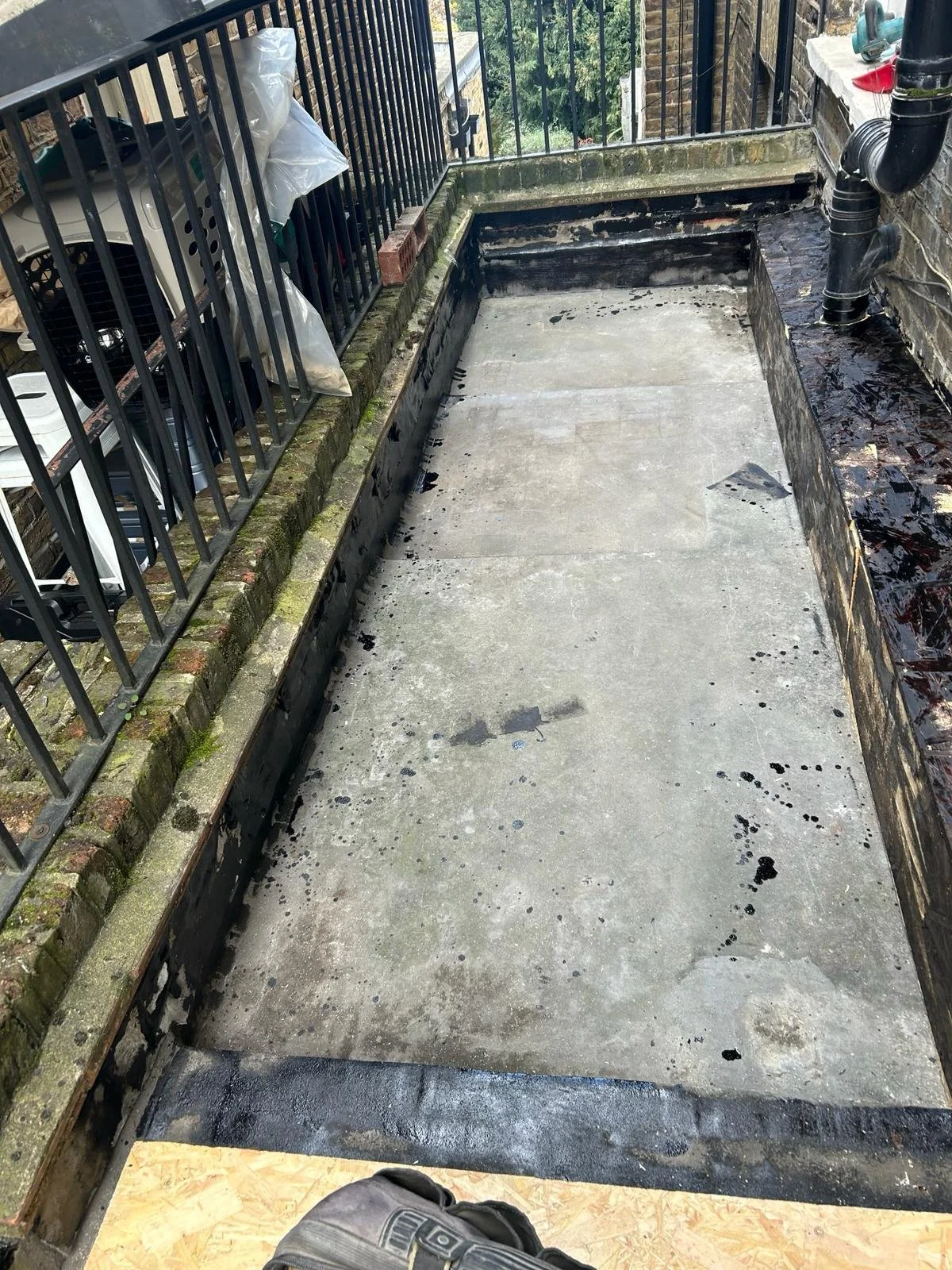 View of a small balcony or rooftop with a concrete surface, black rain gutter pipe, and metal railing; some items and tools on one side and black sealant or paint around the edges.