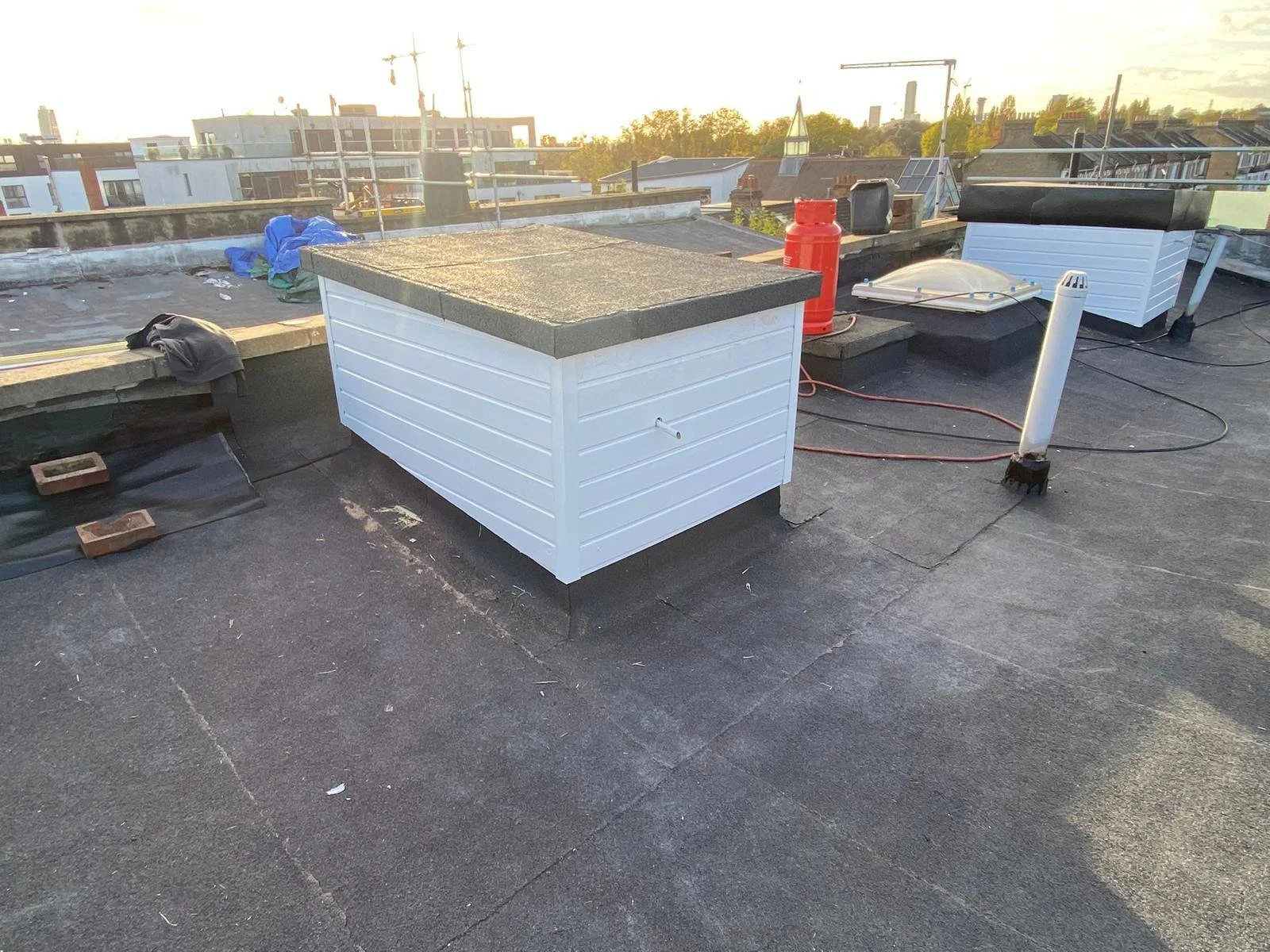 Rooftop with two white vent boxes, a pipe, a red fire extinguisher, and other equipment, with city buildings and factories in the background at sunset.