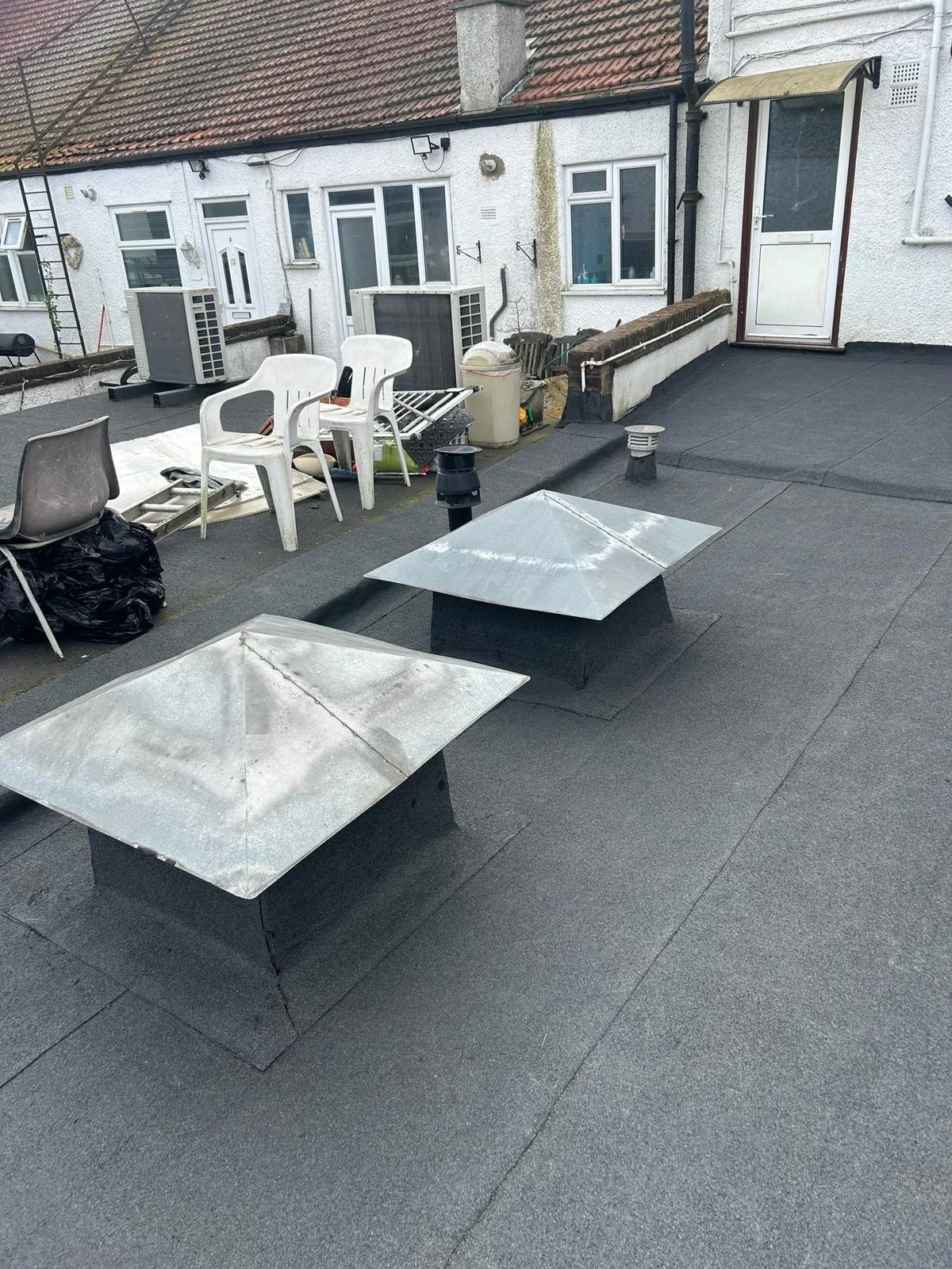 A rooftop with black asphalt covering, two metal vents, and various outdoor furniture and items, including white plastic chairs, a gray chair, a trash bag, and miscellaneous tools and containers. The background shows white residential building walls 