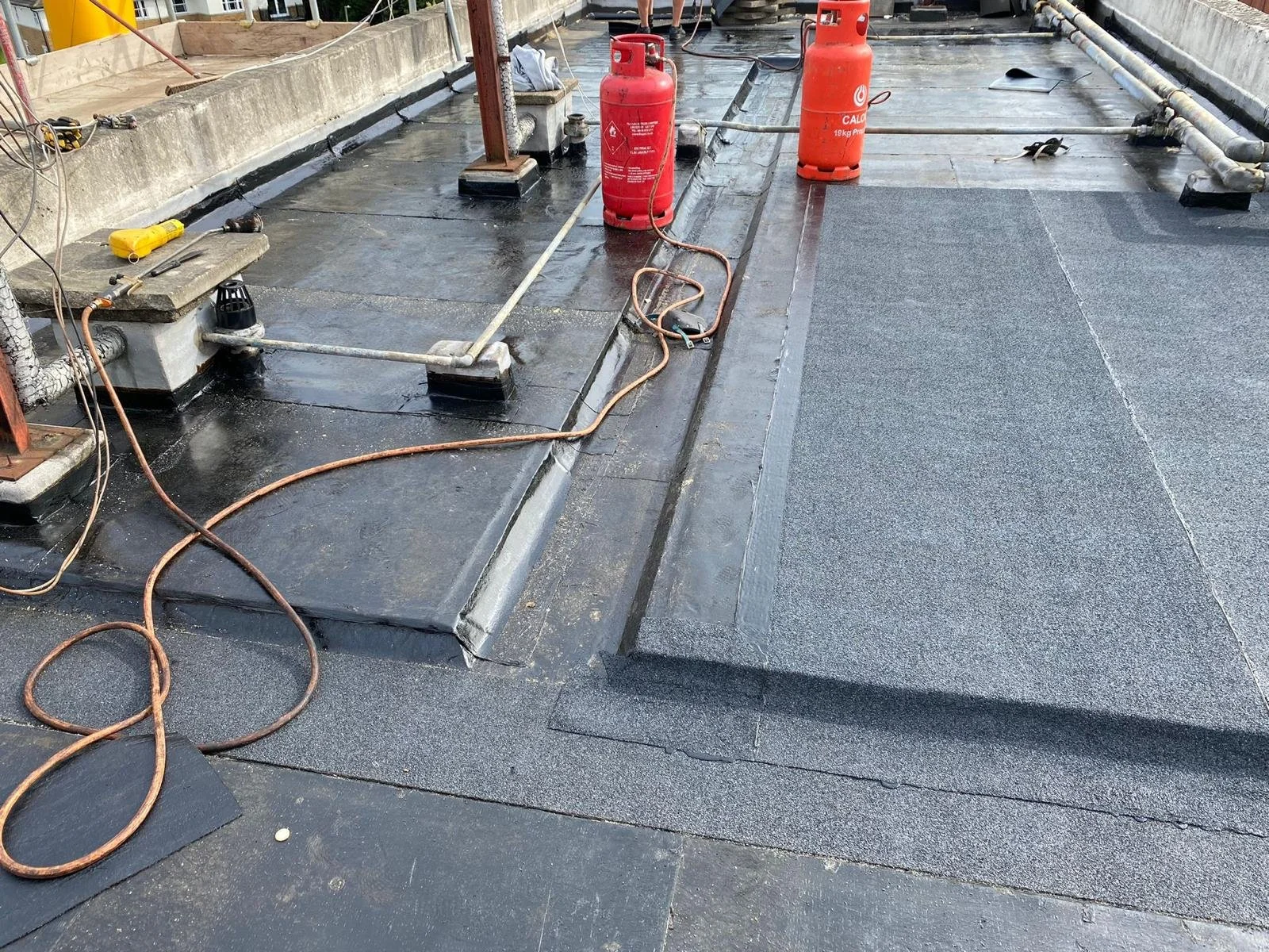 Roof construction site with black waterproofing membrane, orange propane tanks, construction tools, and hoses.