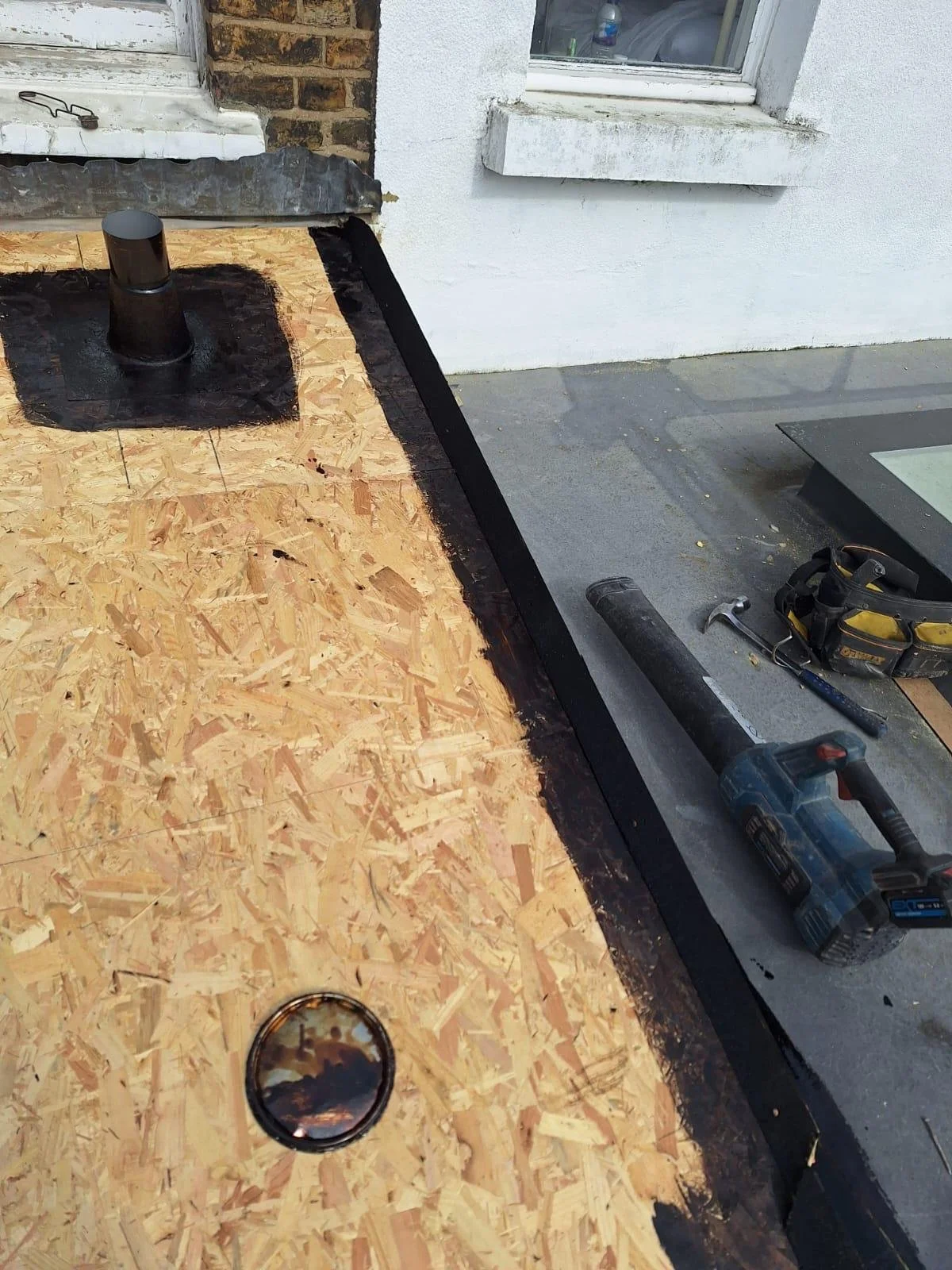 Construction site showing OSB plywood with a skylight and a roof vent, partially covered with black roofing felt, near a white building with a window.