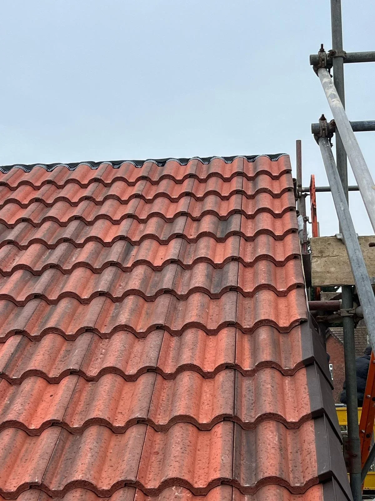A section of a roof with red clay tiles under construction, with scaffolding on the right side and a portion of a ladder visible.