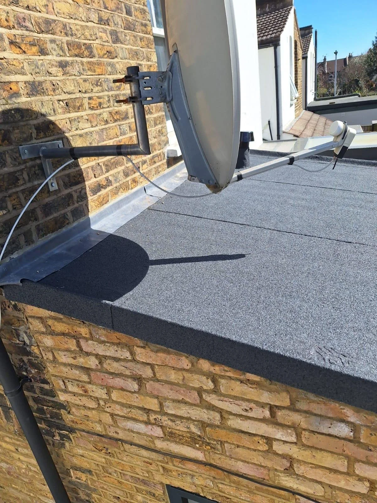 Satellite dish mounted on the roof of a building, with a brick wall and neighboring houses in the background.