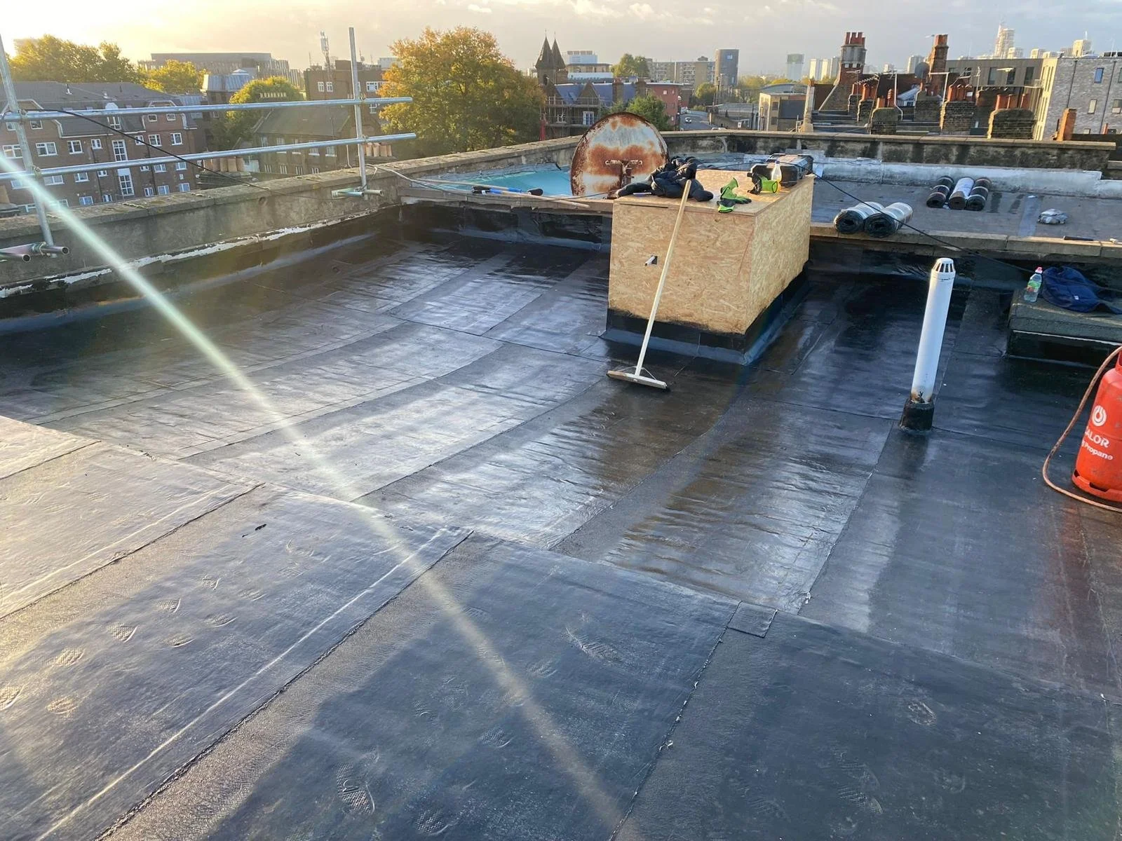 View of a rooftop with black waterproofing membrand, with tools and supplies, city skyline in the background, during sunset.