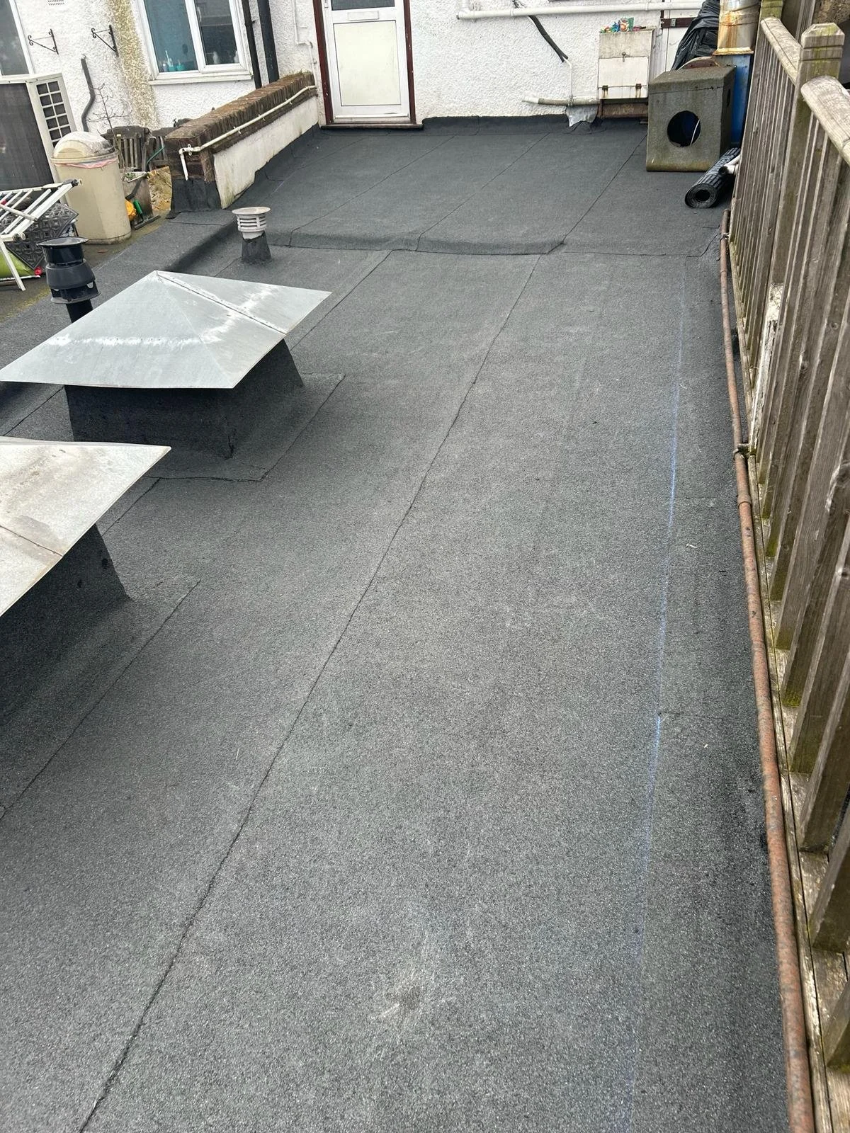 Backyard patio with black rubber roofing material, two metal vents, and a wooden fence on the right side.