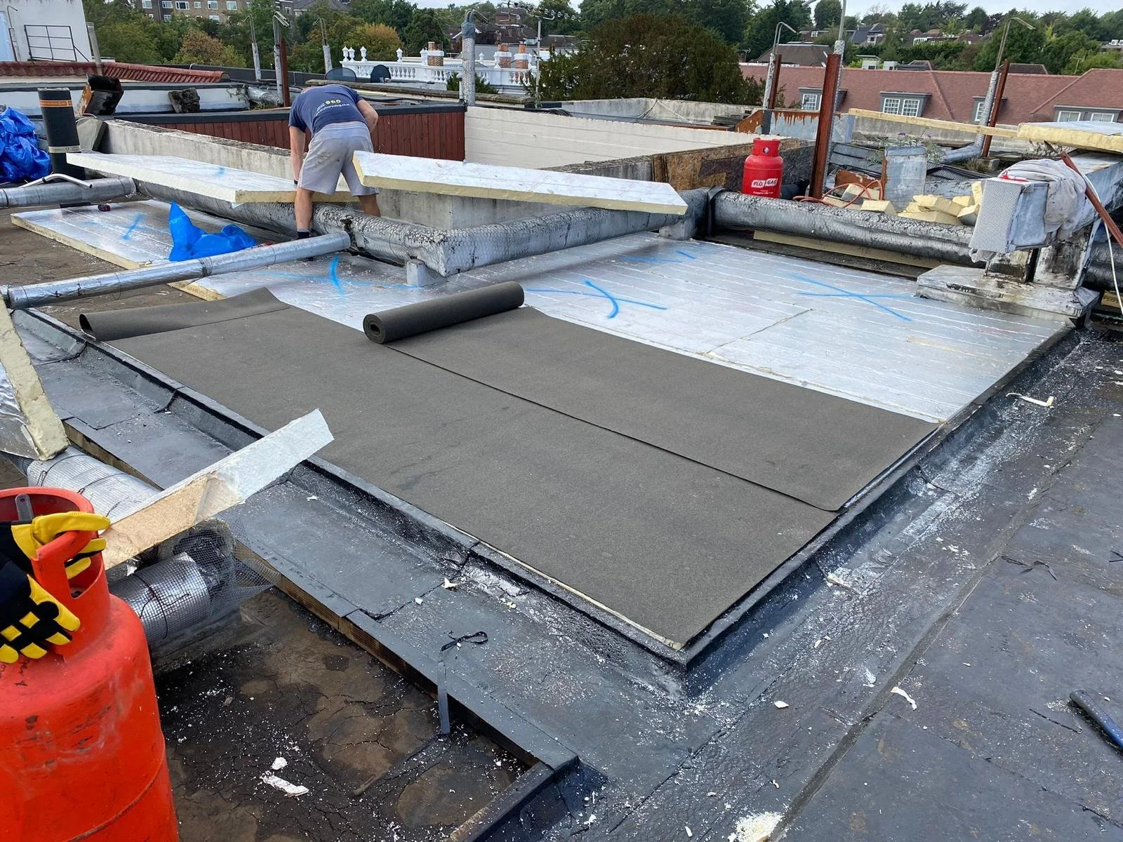 A construction worker installing roofing insulation and heat-reflective material on a building's flat roof while laying down new gray roofing material.