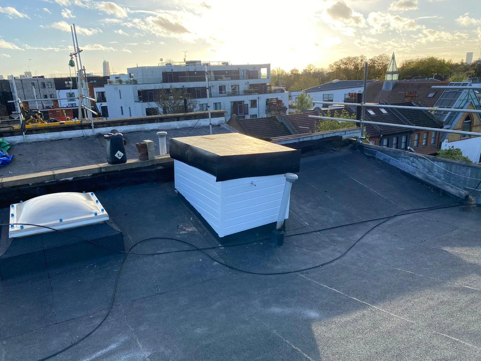 View of a flat rooftop with a skylight and a white vent, overlooking neighboring buildings and trees during sunset.