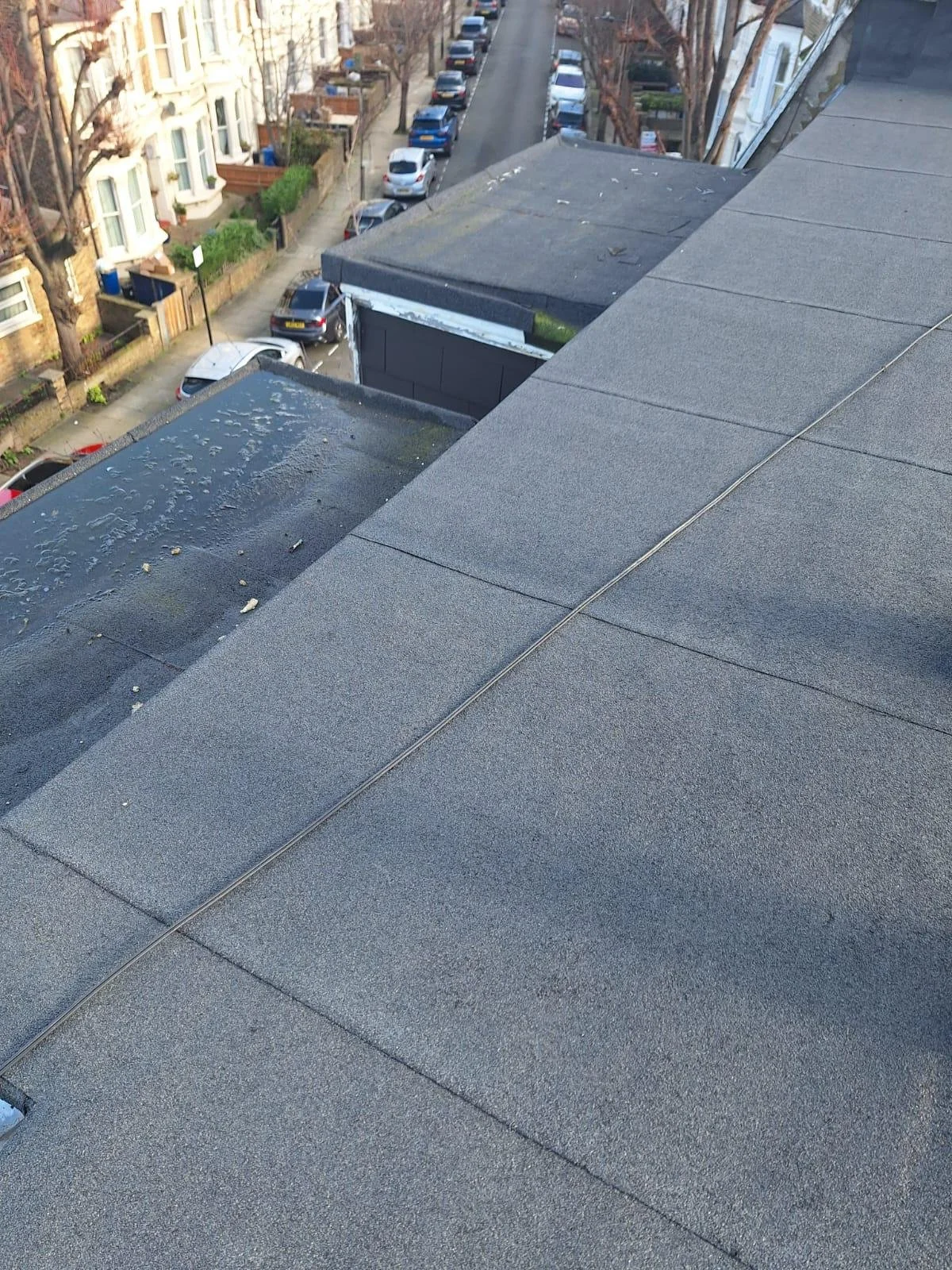 Looking down from a rooftop, showing a street lined with parked cars, apartment buildings, and leafless trees, with a portion of the roof in the foreground covered in gravel or asphalt shingles.